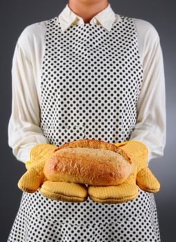 Closeup of a homemaker in an apron and oven mitts holding a fresh baked loaf of bread. Horizontal format over a light to dark background. Woman is unrecognizable. Shallow depth of field.