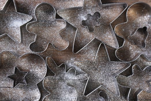 Closeup overhead view of a group of assorted cookie cutters in various shapes on a wood kitchen table sprinkled with flour. Stars, gingerbread men, moose, bell and candy canes are shown. 
