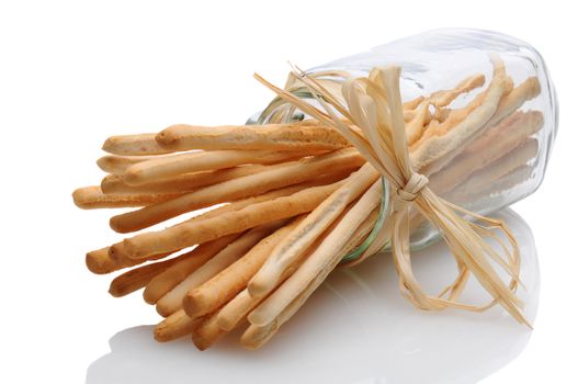 A jar of breadsticks laying on its side on a white reflective surface. The neck of the glass is tied with raffia. Horizontal format.