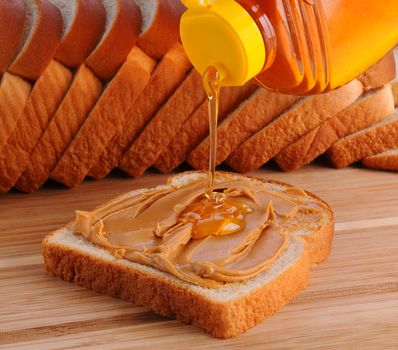 Closeup of making a peanut butter and honey sandwich. Slice of bread with PB  already spread and honey being poured on. Shallow Depth of field with slices of bread in the background.