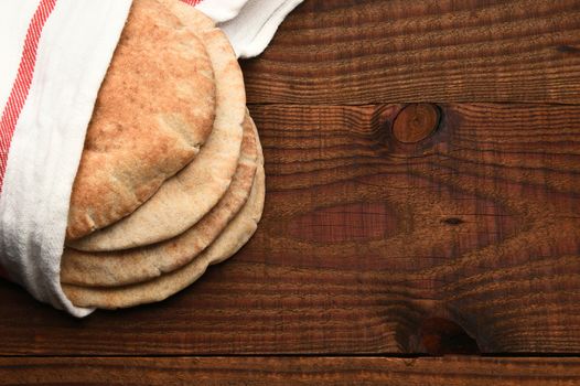 Whole wheat pita bread on a rustic wood table with copy space. The bread is wrapped in a towel and seen from a high angle.
