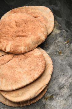 Vertical closeup of loaves of whole wheat pita bread on a gray slate surface and black background.