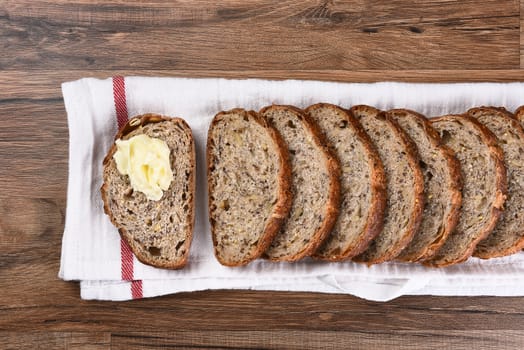 Top view of a sliced loaf of multi-grain bread on a towel, one slice is buttered.