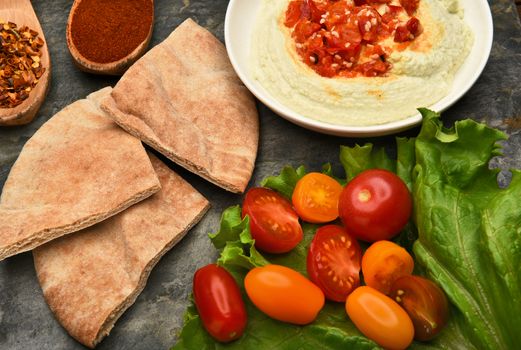 Top view of a bowl of edamame hummus and pita wedges on a slate surface. Lettuce leaf, tomatoes, and spices fill the scene.