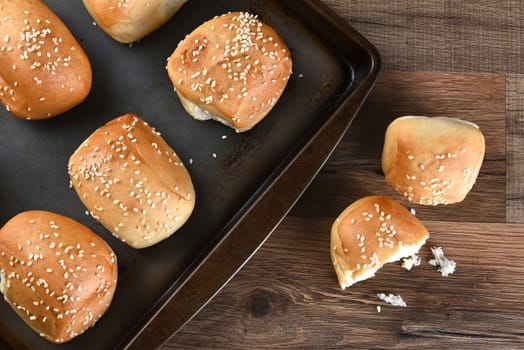 High angle view of fresh baked sesame seed dinner rolls on a baking sheet. A bun broken in half is on the table.