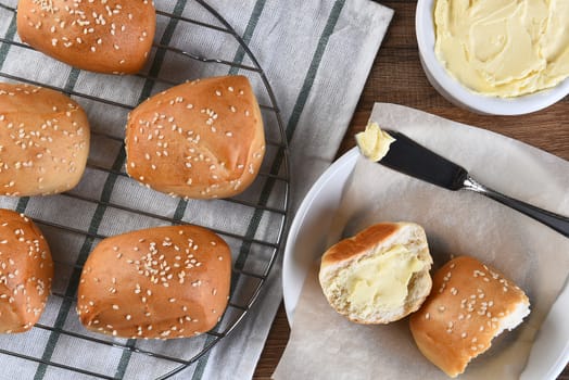 Top view of sesame seed dinner rolls on a towel and cooling rack, next to a plate with single cut roll and butter.