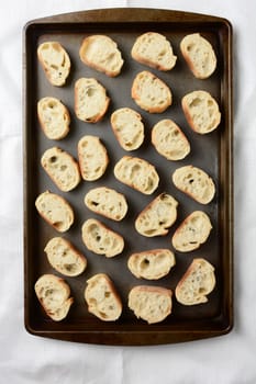 A baguette sliced and spread out on a baking sheet in preparation for toasting. Overhead view in Vertical format.