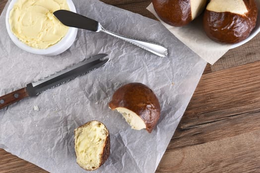 HIgh angle view of a pretzel bun broken in half and buttered. A crock of butter, knife and bowl of whole buns with a broken roll on parchment paper.