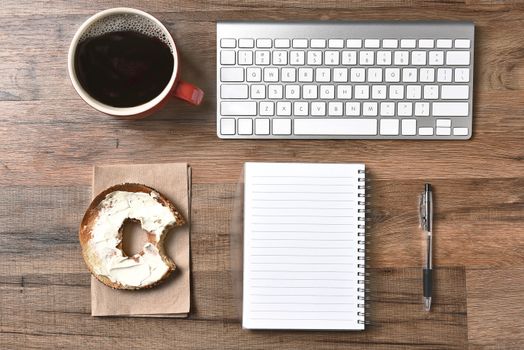 A bagel with cream cheese and a bite taken out next to a computer keyboard and a pad and pen. A cup of coffee in the upper corner, Horizontal for a high angle.