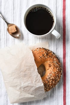 Fresh bagel, hot cup of coffee and spoon with lump of brown sugar on a tea towel. High angle view in vertical format.