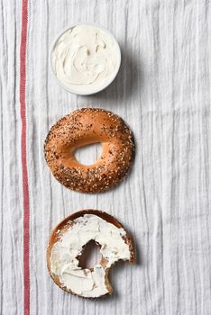 Overhead view of a sliced bagel, one half spread with cream cheese. A crock full of spread on a kitchen towel.