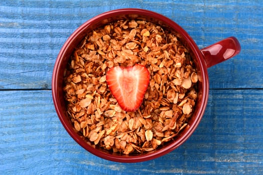 High angle shot of a large mug filled with healthy whole grain cereal with a single sliced strawberry in the middle. Horizontal format on a blue painted rustic kitchen table.