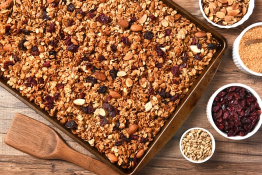 Horizontal overhead view of fresh homemade granola. Closeup of a baking sheet filled with the tasty, healthful food with containers of ingredients.