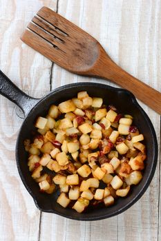 High angle shot of Fried Breakfast Potatoes in a cast iron skillet. Peppers, onions and potato cubes fill the skillet resting on a rustic farmhouse style kitchen table with a wooden fork.