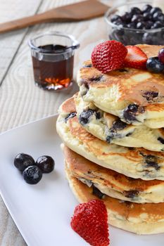 Closeup of a homemade blueberry pancake breakfast with syrup pitcher, wooden spatula, blueberry bowl and strawberries. Vertical format with shallow depth of field.