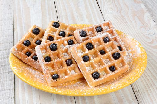 Closeup of a plate of waffles and blueberries. The plate is on a rustic farmhouse style table and the waffles are covered with powdered sugar. Horizontal format.
