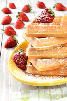 Waffles and strawberries covered with powdered sugar. The stack of waffles is on a yellow platter on a rustic farmhouse style kitchen table. 