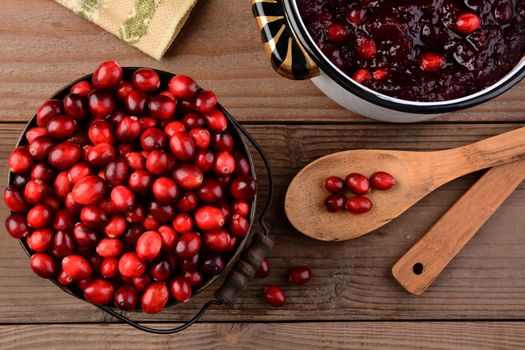 Overhead of a bucket of cranberries and a pot full of whole cranberry sauce on a rustic wooden table. Cranberry sauce is a traditional Thanksgiving side dish. Horizontal format.