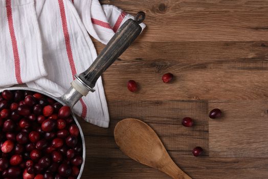 Fresh cranberries in a pot ready for cooking. Scattered berries a spoon and kitchen towel are on the wood table.