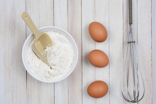 Overhead image of a bowl of flour, three brown eggs and a metal whisk on a rustic wooden kitchen table. 