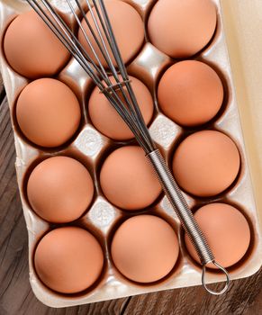 High angle view of a carton of brown eggs with a whisk laying across. The carton is resting on a rustic wooden farmhouse table. 
