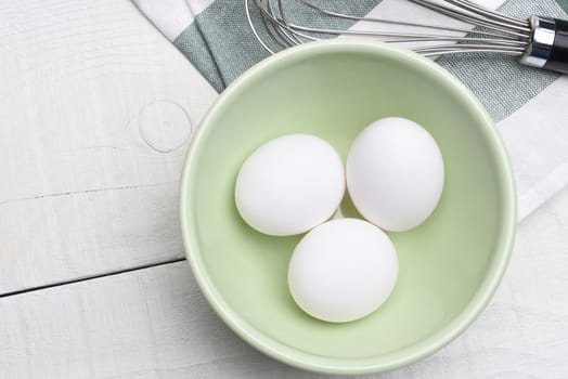 High angle view of three eggs in a bowl on a wood table with kitchen towel and whisk.