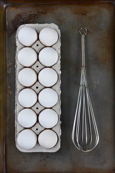 HIgh angle image of a carton of eggs and a wire whisk on a metal baking sheet. Vertical format. 