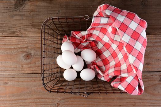 A wire basket with eggs and a red checkered napkin on a rustic wood surface. Overhead shot looking straight down into the basket.