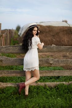 Attractive smiling model girl in a dress and light summer hat with a brim posing in the countryside. Lifestyle summer portrait of a beautiful young woman wearing a hat with brim posing in a rural background