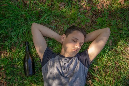 drunk man sleeping on a green field after using alcohol, a bottle lying next to a man.