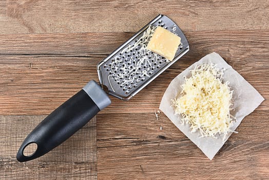 Parmesan Cheese and grater on a wood kitchen table. Some grated cheese is on a piece of parchment paper next to the grater. High angle view in horizontal format.