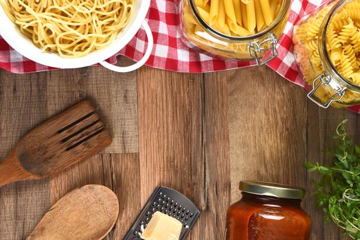 High angle view of an assortment of pastas, parmesan, cheese, grater, jar of sauce, utensil and herbs on a wood kitchen table with copy space.