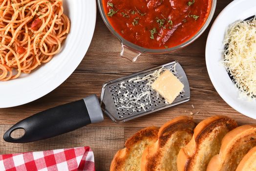 High angle view of an Italian meal on a rustic wood kitchen table. A plate of spaghetti, bowl of sauce, grated parmesan cheese, and garlic bread.