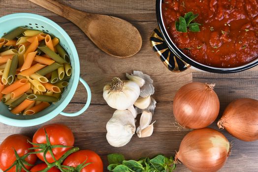 Italian meal ingredients on a wood kitchen table. A pot of sauce, colander of rigatoni, tomatoes, garlic, oregano, and onions seen from a high angle. 