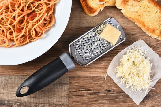 High angle view of a cheese grate with parmesan cheese on a rustic wood kitchen table . A plate of spaghetti and garlic bread are also shown.