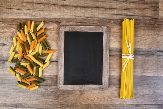 Top view of colorful rigatoni pasta and spaghetti with a blank chalkboard.