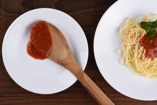 High angle view of a plate of spaghetti with marinara sauce and garnish. A second plate with a wood spoon and sauce in horizontal format.