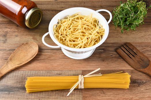 Still life of dried spaghetti, cooked pasta in a colander, a jar of sauce, herbs and wood utensils.