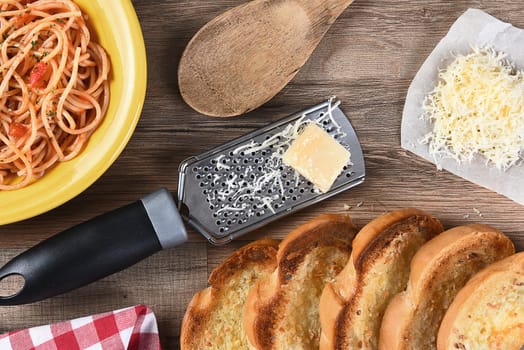 Overhead view of a kitchen table with a plate of spaghetti, garlic bread, grated parmesan cheese, a grater, wood spoon, a red checkered napkin. 