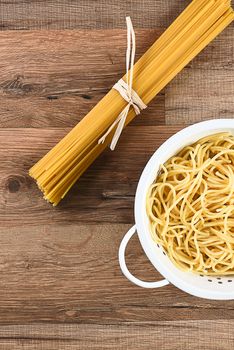 Overhead view of cooked and dried spaghetti. The cooked pasta is in a colander and the raw is tied with raffia. Horizontal on a wood kitchen table.