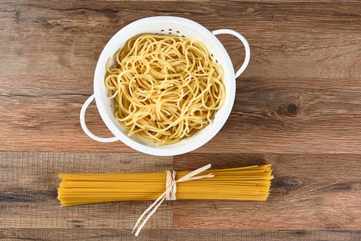 Overhead view of cooked and dried spaghetti. The cooked pasta is in a colander and the raw is tied with raffia. Horizontal on a wood kitchen table.