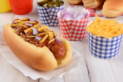 Closeup of a grilled chili dog with cheese and onions on a rustic wood picnic table. More buns and condiments fill the background. 