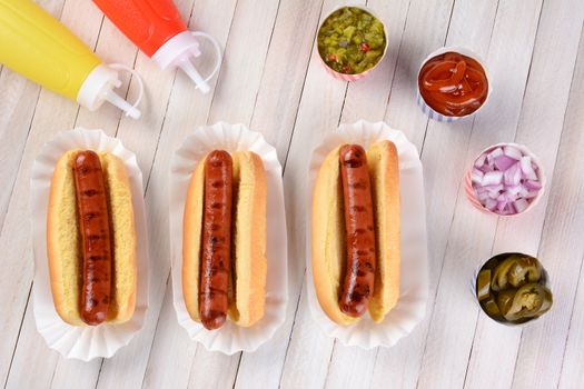 Three grilled hot dogs ready to be garnished on a rustic wood picnic table. High angle view.