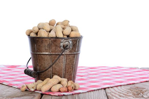 Closeup of a pail full of peanuts on a wood picnic table and a red checked tablecloth. Horizontal format with a white background.