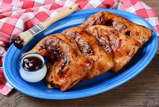 High angle shot of chicken leg quarters on a blue oval platter. The legs have been barbecued and are coated in sauce. Horizontal format on a rustic wooden restaurant table.