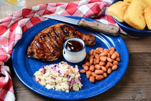 Closeup of a Barbecue chicken plate with cole slaw, pinto beans and corn bread. The meal is on a rustic wooden restaurant table with a red and white checked napkin and a glass of lemonade.