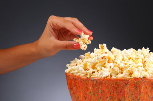 Closeup of a womans hand taking some popcorn from a bowl. Horizontal format over a light to dark gray background.