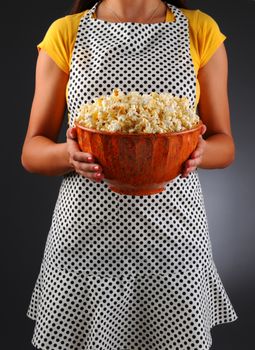 Closeup of a homemaker in an apron holding a bowl of popcorn. Vertical format over a light to dark background. Woman is unrecognizable. Shallow depth of field.