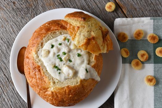 A bread bowl of New England Style Clam Chowder on a rustic wood table with napkin and oyster crackers.