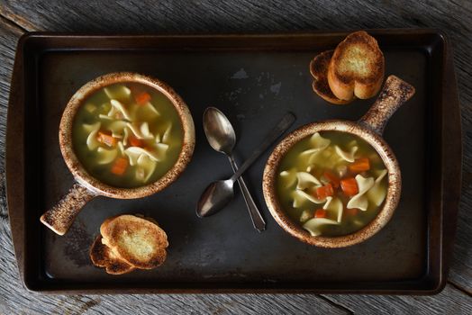 Two bowls of Chicken Noodle Soup, a Baguette  Garlic Toast and spoons on a metal baking sheet on a rustic wood kitchen table.